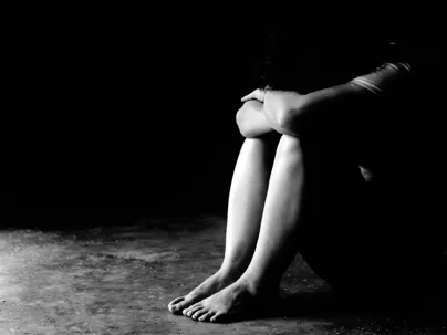 A child sitting in a dark corner, head bowed and covered by their arms and knees, in black and white color.