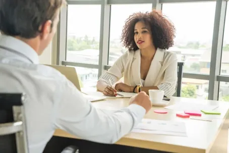 A man and a woman talking face to face, with papers, a cup of coffee, and a personal computer on the table.