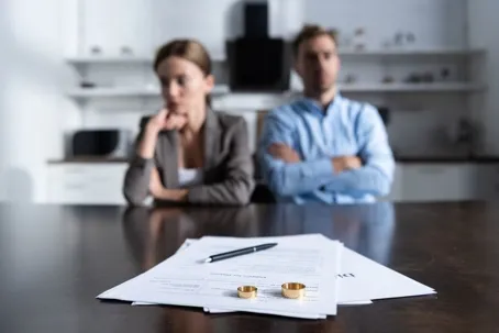 On the table are divorce papers, wedding rings, and a pen, with the couple sitting beside each other.