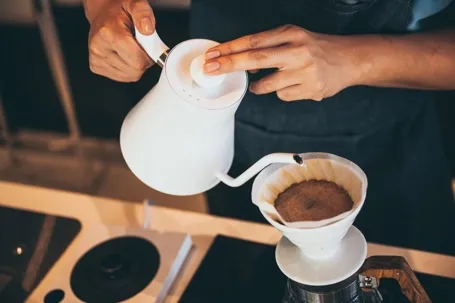 A person pouring hot water from a white kettle into a coffee dripper with a paper filter to make pour-over coffee.