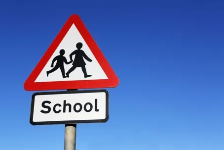 A school crossing warning sign showing two children walking, with the word “School” below, against a clear blue sky.