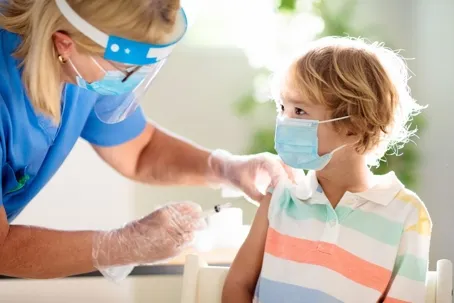 A nurse wearing a face shield and face mask is giving a vaccine to a little boy who is also wearing a face mask.