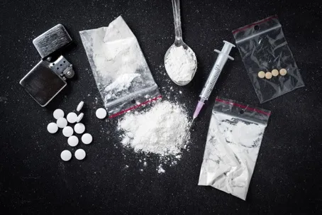 Various illegal drugs and paraphernalia on a dark surface, including white powder, tablets, plastic bags, a syringe, and a spoon.