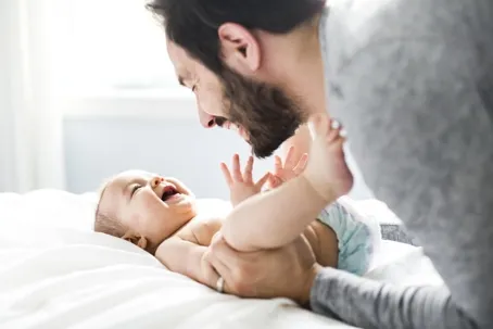 A father and his cute little baby are playing and laughing while the baby lies on the bed facing his father.