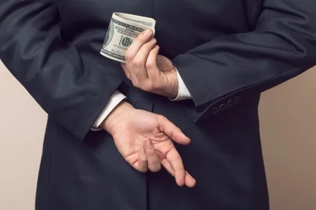 A man in a suit hiding a stack of dollar bills behind his back while crossing his fingers.