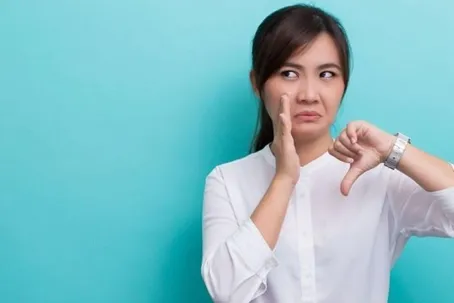 A woman in a white shirt showing a thumbs-down gesture with a displeased facial expression, standing against a blue background.