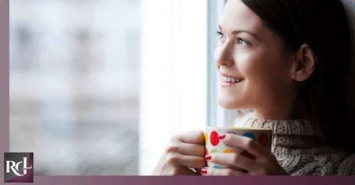 An image of a smiling girl looking out the window while holding a cup.